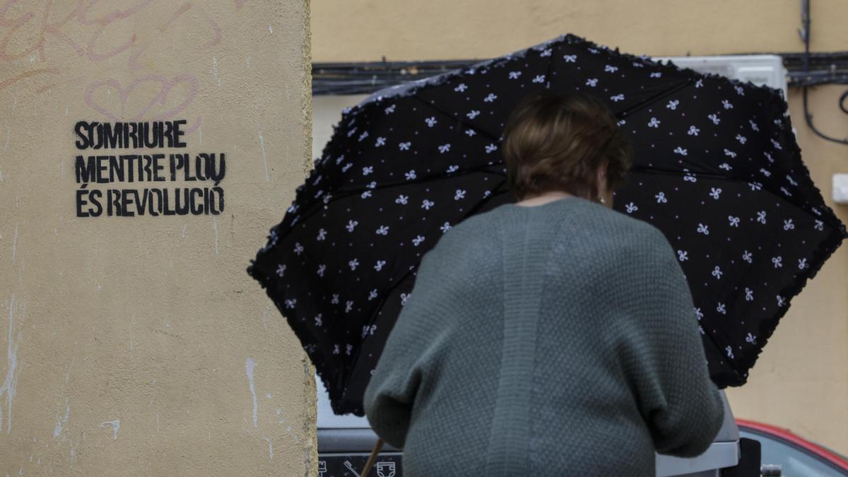 Una mujer se protege de la lluvia con un paraguas cuando pasa junto a una pintada con el lema "Sonreir mientras llueve es revolución".